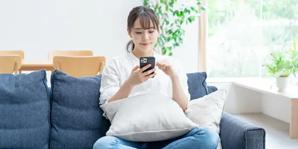 岡山・倉敷のロケーション撮影専門であるスタジオグッドの撮影予約をするためのリンク|ソファに座った女性がスマートフォンで自宅から簡単に予約している様子の写真